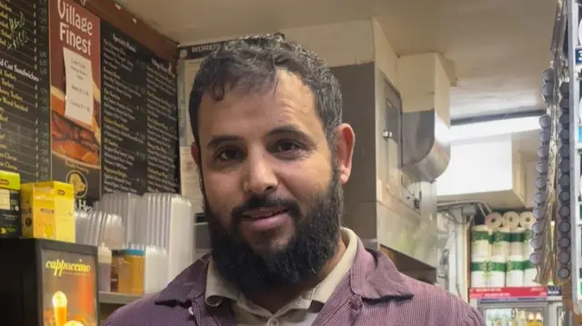 Yousef Hashmi in a deli with a menu board behind him