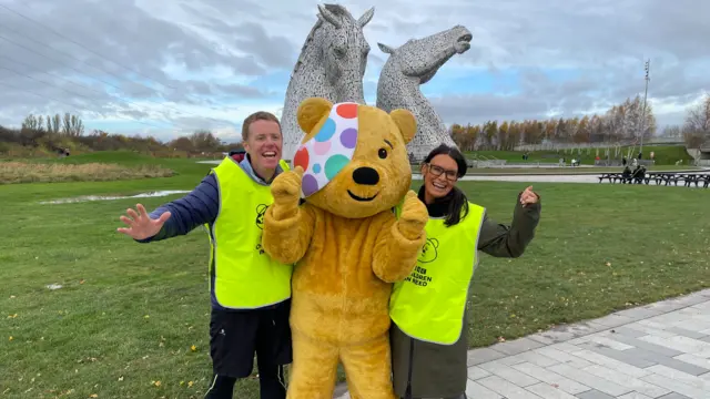 Two people stood next to somebody dressed in a yellow Pudsey bear suit. They are both smiling and holding their arms up to celebrate.