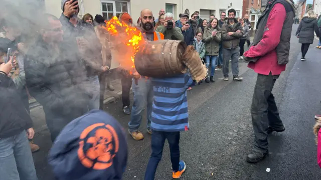 A crowd of people watching as a boy carries a flaming tar barrel above his head.