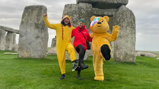 A man and woman pictured in front of Stonehenge. They are stood with somebody dressed up as Pudsey Bear.