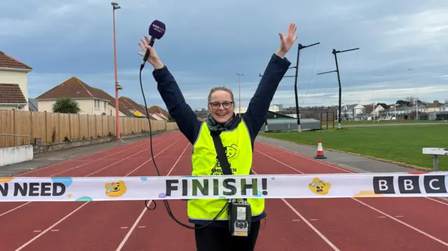Jenny Mullin stands with her hands in the air, a purple BBC Radio Jersey mic in her left hand. She is wearing a high vis Children in Need bib and standing on a red racing track in front of a finish line which has pictures of Pudsey's face on and says 'Finish!'