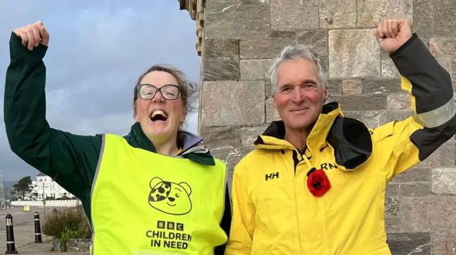 Caroline Densley from BBC Radio Devon in a yellow Children in Need tshirt with a RNLI crew member in yellow uniiform cheering next to a monument