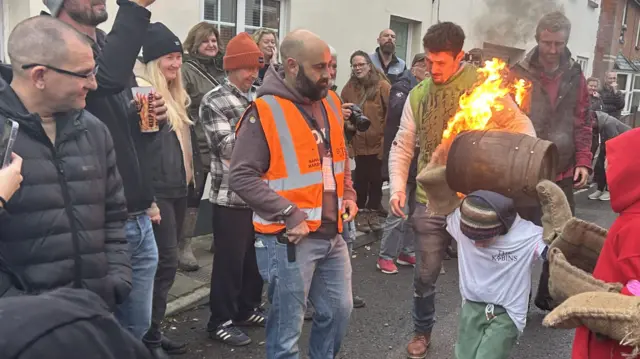 A young boy with a tar barrel on his head as crowds watch on.