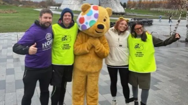 Two men and two women stood in a line next to somebody in a Pudsey costume. They are pictured outside. They are all smiling.
