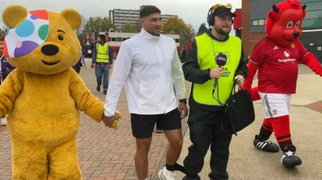 Pudsey Bear holds Tommy Fury's hand, Tommy is wearing a white jacket and black shorts, his leg is tied to Joe McGrath's. Joe has headphones, a purple mic and yellow high vis Children in Need bib. A Manchester United Fred the Red Devil mascot walks beside him.