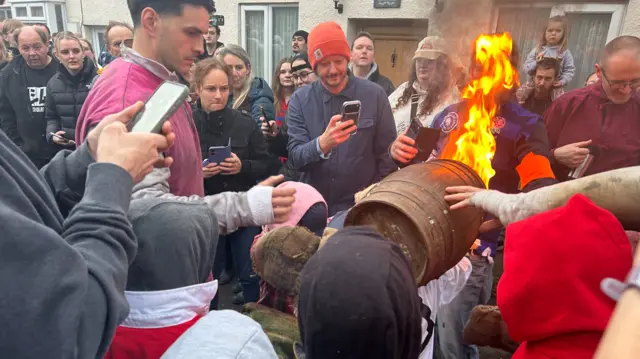 A crowd watching on as a young person carrying a flaming tar barrel above their head.