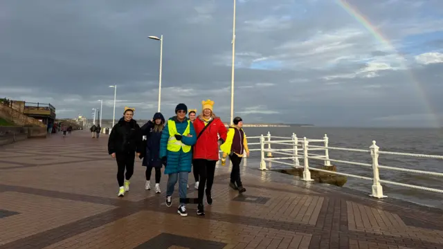 Several people walking along a seafront. There is a rainbow in the sky above them