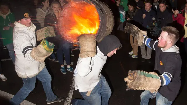 Three men are seen with hessian bags around their hands and a flaming tar barrel on their back.