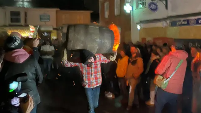Crowds gathering to watch a person carrying flaming tar barrel .