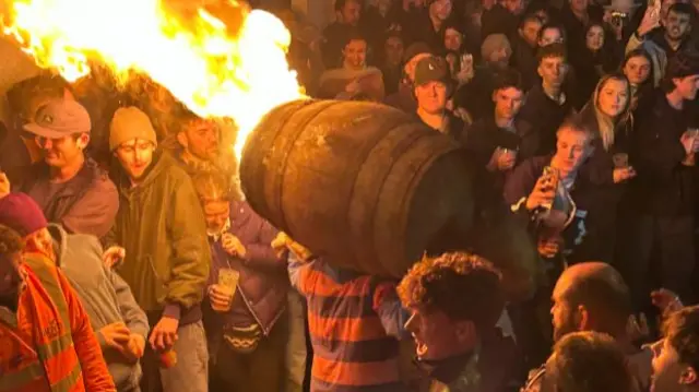 A person running with a flaming tar barrel on his back.