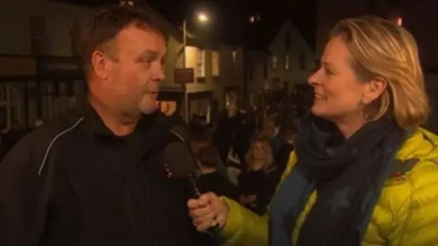 Alex Rummery in a black jacket speaking to Victoria Graham, in a mustard jacket.