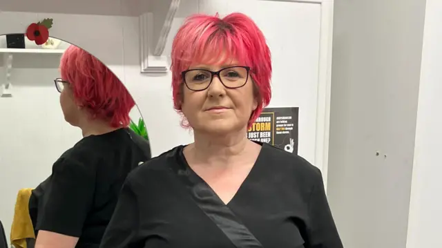 Petra Brown leaning against one of the tables in her barber shop. She has red hair, is wearing glasses, and a black top.