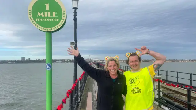 A woman and a man on a pier. The woman has blonde hair and is wearing yellow Pudsey bear ears. The man is also wearing the ears and has a yellow high vis Children in Need vest on. Both of them have their arms up to celebrate and are smiling at the camera.