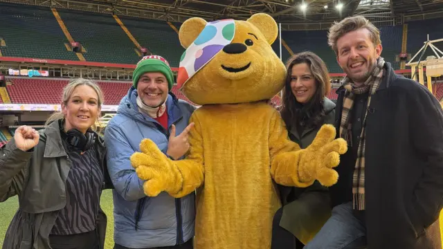 Several people and Pudsey Bear stand in front of stands at a sports stadium