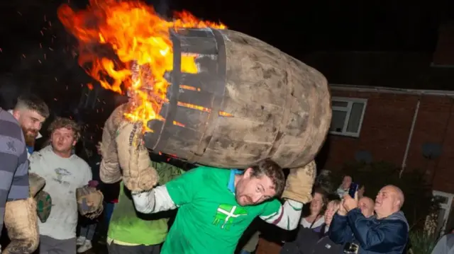 A man in a green T-shirt carrying a barrel, which is on fire, as people watch on
