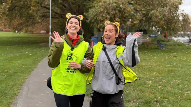 Presenter Katie Tyler and journalist Ellie Tutt wearing Pudsey ears and Children in Need bibs waving on a path alongside the Thames