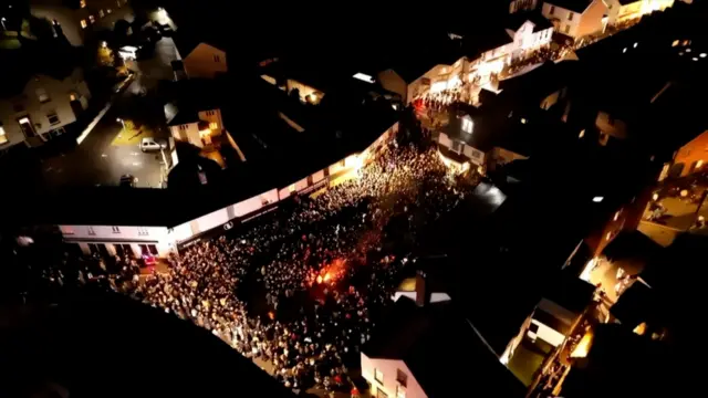 A drone shot of Ottery St Mary at night. There is a burning tar barrel at the centre of a crowd of people.