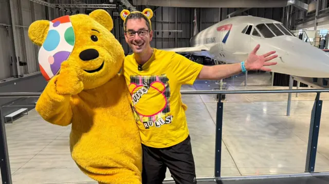 Somebody dressed up as Pudsey Bear and standing next to a man with short dark hair and glasses. They are pictured in Aerospace Bristol. There is a concorde plane in the background behind them.