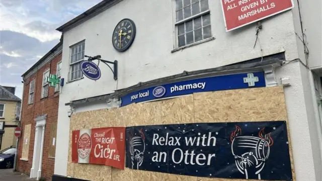 A pharmacy boarded up with signs across the front. There is a clock above the brown boards which have been placed across the windows.
