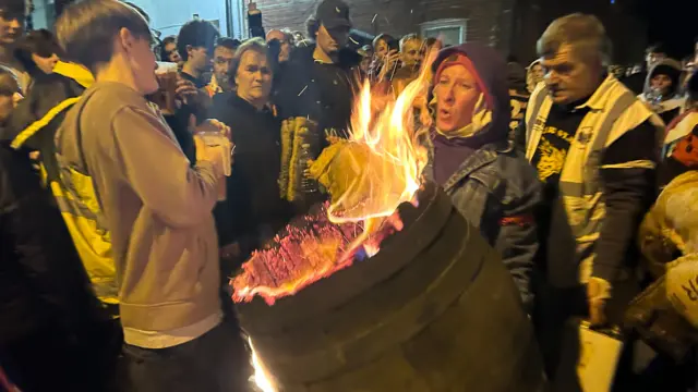 A crowd gathers around a flaming tar barrel.