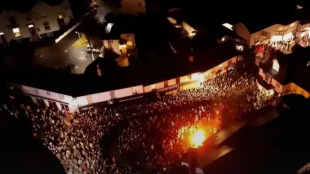 A drone image of crowds and there is a flaming barrel in the middle of it.