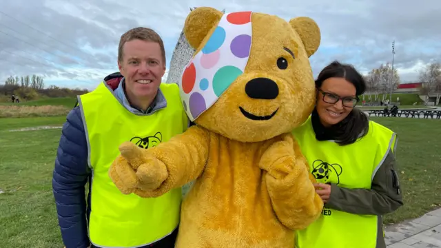 A man and a woman with somebody dressed up as Pudsey bear. The man has short fair hair and the woman has dark long hair. They are both looking at the camera and smiling