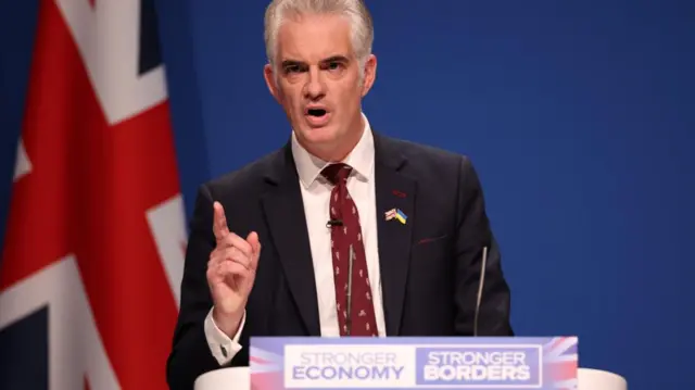 South Suffolk MP James Cartlidge speaks from a lectern and raises a finger.