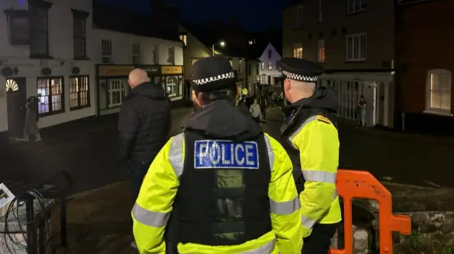 Two police officers stood at the top of a hill watching the crowd. They are wearing yellow hi-vis jackets with the words 'Police' on the back. It is dark and the street is lit with street lights.