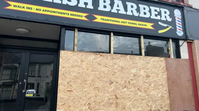 A barber shop with a boarded up window on the front. The board is light brown and the door is not covered.