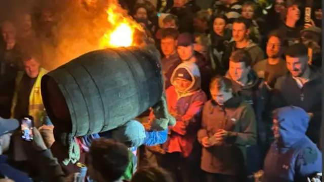 A person running with a flaming tar barrel on his back.