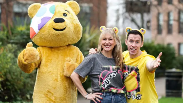 Somebody dressed in a yellow Pudsey bear suit standing next to a woman and a man. The woman is blonde and is wearing yellow Pudsey ears. The man has dark short hair and is also wearing Pudsey ears. They are both looking at the camera and smiling.