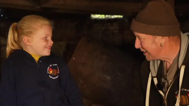 Jessica is sitting on a brown wooden barrel. She has tied-back blonde hair, is smiling, and wearing a navy fleece. Les is wearing a brown woolly hate and a brown hoodie.