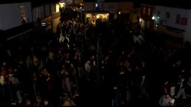 A crowd of people in a dark street with the glow from buildings behind.