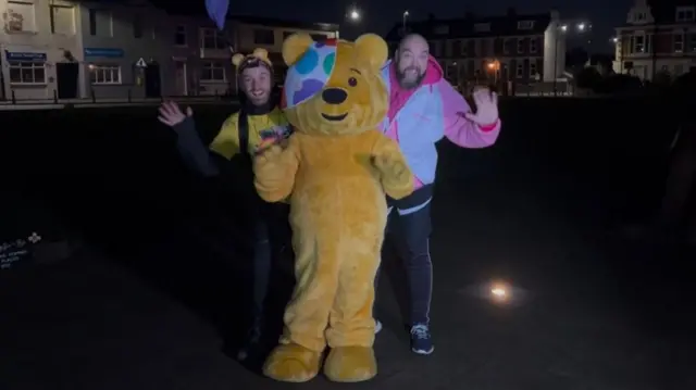 Matt Bailey, Pudsey and Colin Bergman Plews are waving at the camera as they stand in the dark outside Soldier 1101.