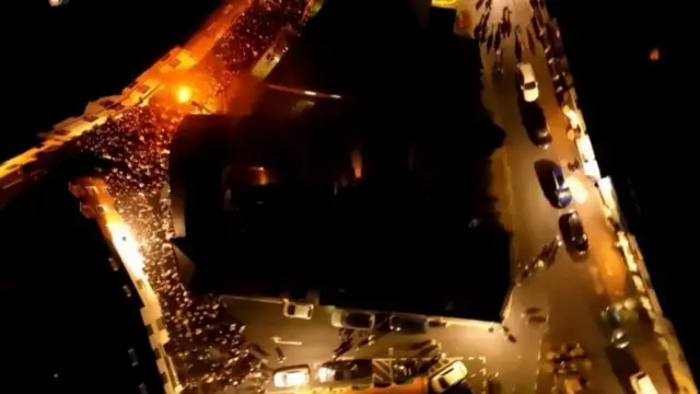 A drone image of crowds and there is a flaming barrel in the middle of it.