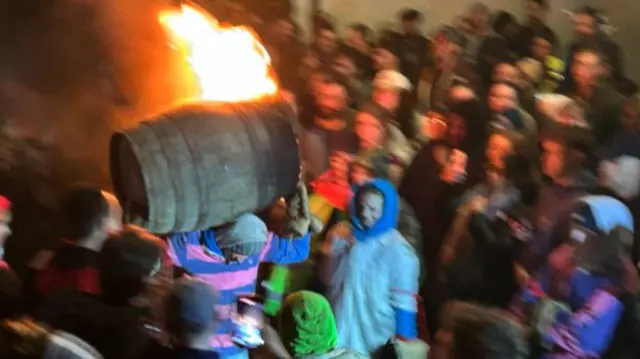 A person running with a flaming tar barrel on his back.