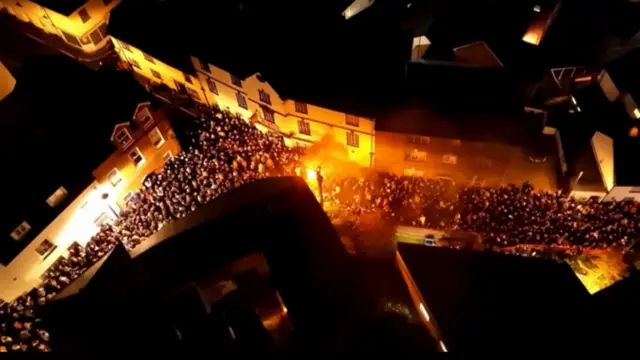 Drone footage of a lit barrel glowing amongst a crowd of people in the streets below. It is dark and the roofs of buildings can be seen.