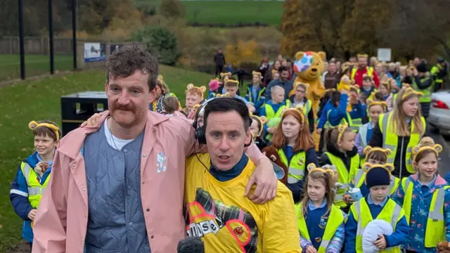 Two men walking in front of a large crowd of primary school children. They have their arms around each other and are looking straight ahead.