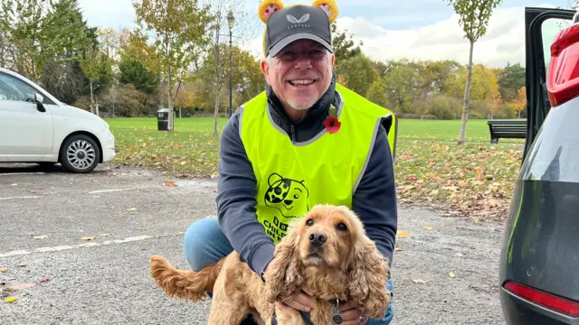 A man crouched down to stroke a brown dog. He has white stubble on his chin and is wearing a yellow high vis vest and yellow Pudsey ears. He is looking at the camera and smiling.