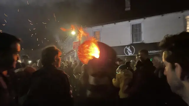 Crowds gathering to watch a person carrying flaming tar barrel .