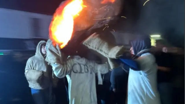 People helping carry a flaming tar barrel. They are wearing hessian gloves.