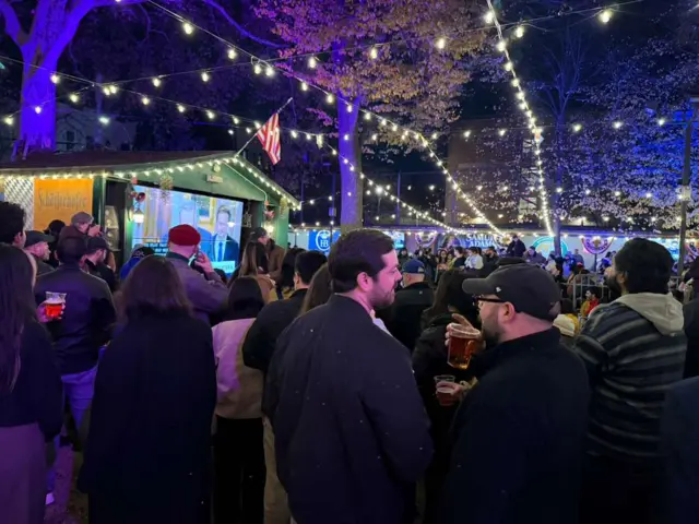 Several people stand in front of a large TV outdoors, under twinkle lights at an election night watch party in New York City