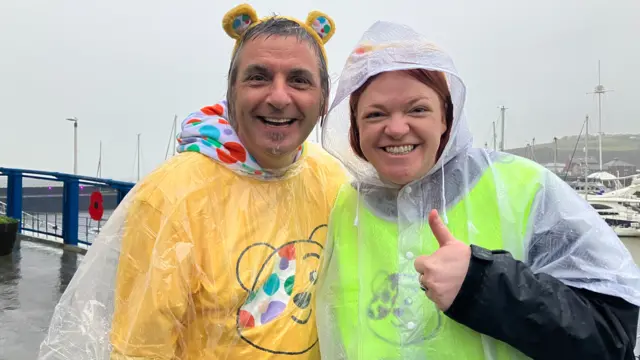 A man and a woman pictured outside in rain ponchos. They are both looking at the camera and smiling.