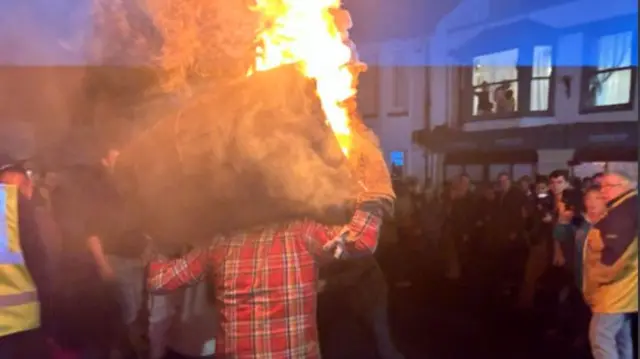 A person wearing a red checked shirt carrying a flaming barrel above his head. There is a crowd of people watching them.le