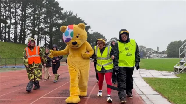 A group of people walking on an orange running track. Two of the people are wearing high vis jackets and have one leg tied for a three-legged race. Somebody dressed in a yellow Pudsey bear suit is walking alongside them.
