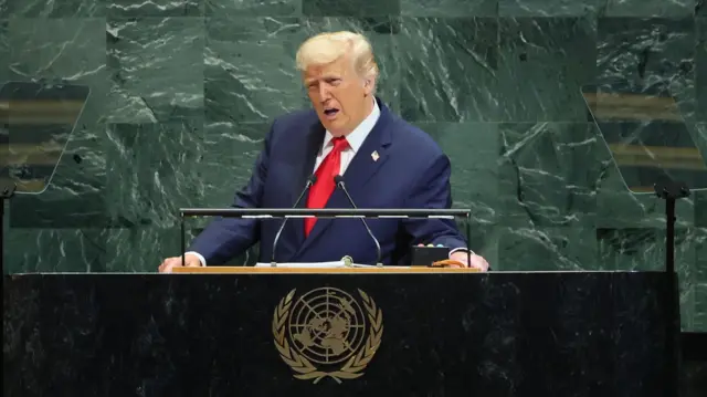 President Trump wears a blue suit and red tie. He stands in front of a black podium with the UN logo emblazoned in gold. Behind him is a green marble wall