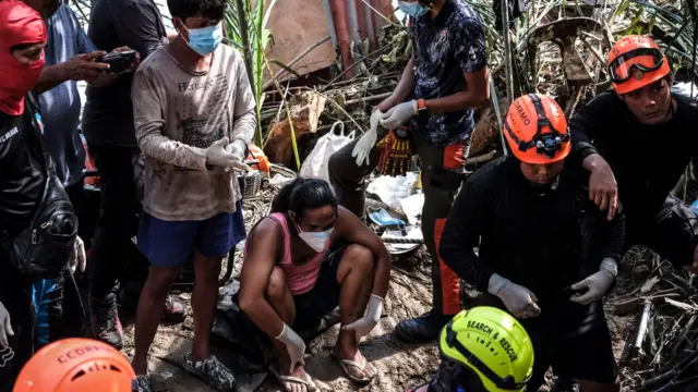 A group of emergecny responders wearing orange and yellow hard hats are working. Civilians stand and sit, wearing masks.