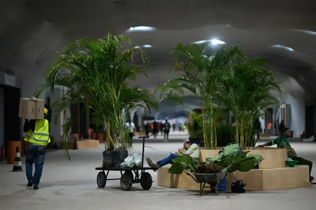 Long corridor in the COP30 venue, beige carpet and domed ceiling. Worker can be seen napping on a bench and tree pots around her. A construction worker in a hi-vis jacket walks with a box on his shoulder to the left
