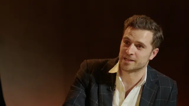 Alexander is wearing a white shirt and a checkered black blazer.