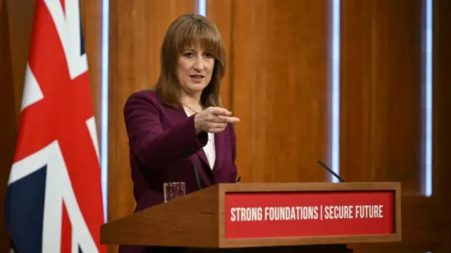Reeves points with one hand as she delivers a speech. She stands at a lectern, on which the words 'strong foundations, secure future' are printed. She is flanked by a furled union flag